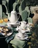A table set for breakfast outdoors, with a teapot, cups, pastries, and orange juice, set against a backdrop of large cacti. Náhľad obrázku produktu