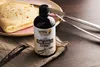 A bottle of vanilla extract is placed next to a folded crepe on a napkin, with vanilla beans in the foreground and cutlery in the background. Produktbild-Vorschau