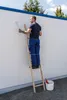 A man is painting a white wall while standing on a wooden ladder. A bucket with a red object is on the ground next to the ladder. Produktbild-Vorschau