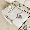 A modern white coffee table with gold accents is placed on a patterned rug in a living room setting, next to a white sofa. Produktbild-Vorschau