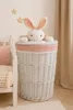 A white wicker laundry basket with a pink gingham trim and a plush bunny toy peeking out from the top, placed next to a child's bed. Podgląd zdjęcia produktu