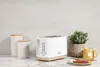 A white toaster with a wooden base is displayed on a marble countertop, alongside two canisters, a potted plant, and a mug of tea. Produktbild-Vorschau