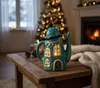 A teal teapot-shaped lantern with illuminated windows sits on a wooden table in front of a decorated Christmas tree and a fireplace. Produktbild-Vorschau