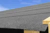 A close-up view of a gray asphalt shingle roof with a clear blue sky in the background and wooden beams on the right side. Produktbild-Vorschau