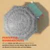 A gray, fuzzy umbrella is shown from above, casting a shadow on a sandy surface. A striped towel and a small ball are near the umbrella. Produktbild-Vorschau