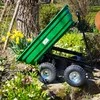 A green garden cart with a tilted bed is shown outdoors in a garden setting with flowers and trees. Náhled obrázku produktu