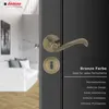 A close-up of a bronze door handle and lock mechanism on a dark gray door, with a blurred interior scene in the background. Produktbild-Vorschau