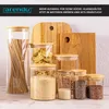 A collection of glass storage jars with bamboo lids, filled with various food items like pasta, grains, and seeds, arranged on a countertop with wooden cutting boards in the background. Náhled obrázku produktu