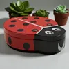 A ladybug-shaped tin box is placed on a light gray surface, with potted succulents in the background. Produktbild-Vorschau