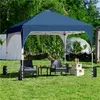 A navy blue pop-up canopy tent is set up in a grassy backyard, with two chairs and a small table placed underneath. In the background, there are trees, bushes, and a geodesic dome structure. Produktbild-Vorschau