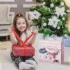 A young girl is sitting on the floor in front of a Christmas tree, opening a gift. A knitting machine box is next to her. Náhled obrázku produktu