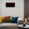 A digital clock with a calendar and temperature display mounted on a wall above a sofa with decorative pillows. Produktbild-Vorschau