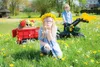 Two children in a grassy field with dandelions, one child in the foreground blowing dandelion seeds, and a red trailer and green toy tractor in the background. Produktbild-Vorschau