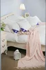 A neatly made bed with white bedding, several white pillows with purple accents, and a pink knitted throw blanket draped over it. A bedside table with a lamp and a decorative lantern on the floor complete the scene. Produktbild-Vorschau