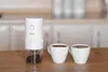 A white electric coffee grinder with a clear container showing ground coffee sits on a wooden table next to two white coffee cups filled with coffee. The background is a blurred kitchen setting. Podgląd zdjęcia produktu
