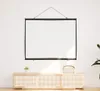 A projection screen is mounted on a white wall above a wooden cabinet with a small plant and a woven box on top. Produktbild-Vorschau