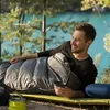 A man is lying in a sleeping bag outdoors next to a body of water. Produktbild-Vorschau