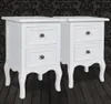 Two white MDF nightstands with two drawers each, placed on a dark wooden floor against a black damask wallpaper. Náhled obrázku produktu