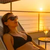 A woman relaxing on a lounge chair by the sea at sunset with a glass of white wine. Produktbild-Vorschau