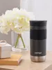 A Contigo travel mug sits on a wooden table next to a small gift box and a stack of books, with a bouquet of white flowers in the background. Náhľad obrázku produktu