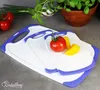 Three white cutting boards with purple trim are stacked on a gray surface. Tomatoes, green onions, and a potted plant are in the background. Anteprima dell’immagine prodotto