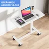 A white adjustable height C-shaped table with a laptop, notebook, phone, glasses, and a coffee cup on it, placed on a wooden floor next to a white wall. Produktbild-Vorschau