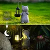 A solar-powered cat statue with a lantern in a garden setting, shown during the day and at night. Produktbild-Vorschau