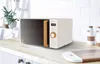 A modern white microwave with a wood-effect handle and orange accents is displayed on a light wood countertop in a kitchen setting. The background features white subway tiles and a ladder with plants. Anteprima dell’immagine prodotto