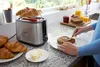A kitchen counter scene featuring a toaster with croissants, butter, toast, jam, and orange juice. Produktbild-Vorschau