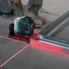 A person's legs and feet are visible in the background, wearing work boots and camouflage pants. A laser level is placed on a concrete floor, projecting red laser lines onto the floor and a metal framing piece. The scene appears to be a construction or ren Produktbild-Vorschau