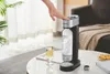 A hand is pressing the top of a Philips soda maker, with a glass of sparkling water and cookies nearby. Podgląd zdjęcia produktu