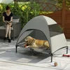 A woman reads a book while sitting on a chair next to a dog resting on an elevated outdoor dog bed with a canopy. Náhľad obrázku produktu