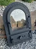 A black cast iron oven door with a glass window, placed outdoors on gravel and rocks. Náhľad obrázku produktu