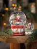 Two Christmas-themed snow globes, one larger with a nutcracker and a smaller one with a teddy bear, are displayed on a wooden surface with pine branches and blurred festive lights in the background. Produktbild-Vorschau