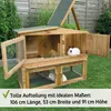 A wooden rabbit hutch with two levels, set up on a grassy lawn with a fence and plants in the background. Two rabbits are visible inside the hutch. Produktbild-Vorschau