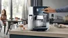 A woman is sitting at a table in a modern kitchen while a Siemens coffee machine is in the foreground. Produktbild-Vorschau