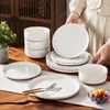 A person is arranging a set of white ceramic dishes on a table runner, with a plant in a pot and a wooden cabinet in the background. Produktbild-Vorschau