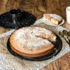 A cake with a slice removed, placed on a decorative cloth on a wooden table. A cake mold and spoons are also visible. Anteprima dell’immagine prodotto