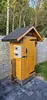 A wooden smoker with a black roof and a metal base, placed on a paved area in a garden. Anteprima dell’immagine prodotto