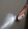 A hand holding a white LED safety light, illuminating a textured gray wall. Produktbild-Vorschau