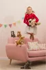 A young girl is jumping in the air, holding a stuffed cat, with several other stuffed cats on a pink sofa. Voir les images du produit