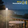 An outdoor floor lamp is illuminated on a patio during a rain shower, with a table and chairs nearby. Podgląd zdjęcia produktu