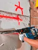 A person wearing a yellow hard hat and safety gloves is using a blue and black Bosch rotary hammer drill to break through a concrete wall. Red spray paint marks are visible on the wall. Produktbild-Vorschau
