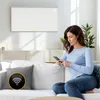 A woman is sitting on a couch and using her phone, with a white infrared heater mounted on the wall behind her. Náhľad obrázku produktu