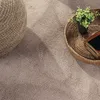 A close-up view of a textured rug with a woven basket containing a small plant and a book on it, and a round woven ottoman in the corner. Produktbild-Vorschau