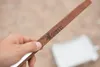 A hand holding a wooden tong with a logo and text on it, with a blurred white object in the background. Produktbild-Vorschau