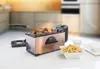 A deep fryer is shown on a white table, with a kitchen background blurred. There are french fries in a bowl and some food being fried in the fryer. Produktbild-Vorschau