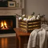 A wooden crate filled with cotton balls and eucalyptus branches, decorated with fairy lights, is placed on a wooden table next to a fireplace with a burning fire. A knitted blanket is draped over the table. Anteprima dell’immagine prodotto
