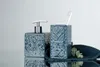 A blue ceramic soap dispenser and toothbrush holder with a toothbrush are displayed on a reflective surface with a dark blue towel and a sprig of greenery. Náhled obrázku produktu