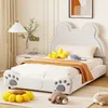 A child's bed with a bear-shaped headboard and paw print details on the footboard, decorated with stuffed animals and a floor lamp. Produktbild-Vorschau